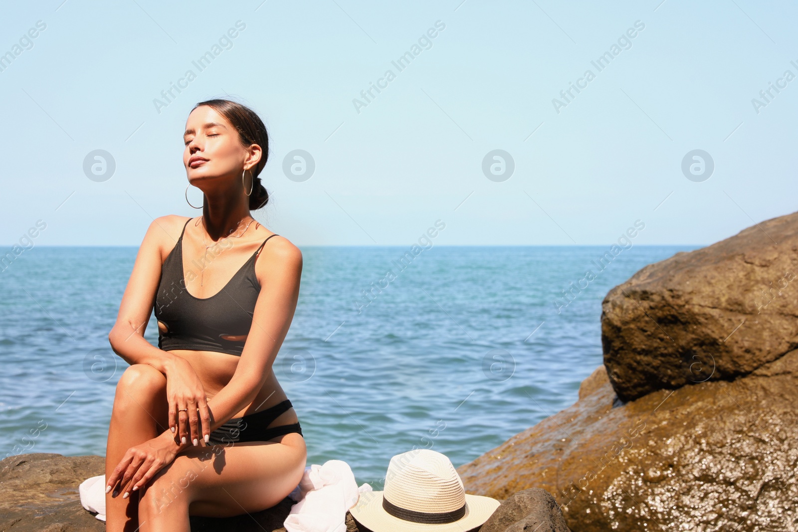 Photo of Beautiful young woman in stylish bikini sitting on rock near sea, space for text