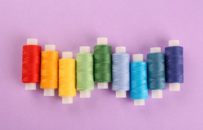 Photo of Different colorful sewing threads on lilac background, flat lay