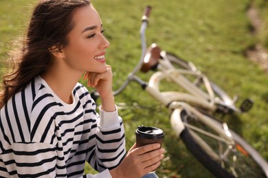 Young woman sitting on green grass and holding cup of coffee near bicycle outdoors, space for text
