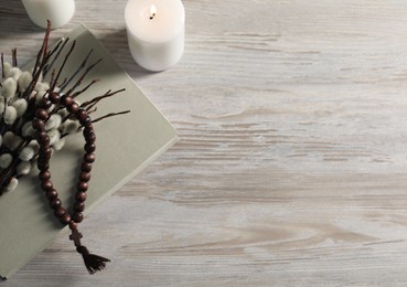 Rosary beads, burning candles, book and willow branches on wooden table, flat lay. Space for text