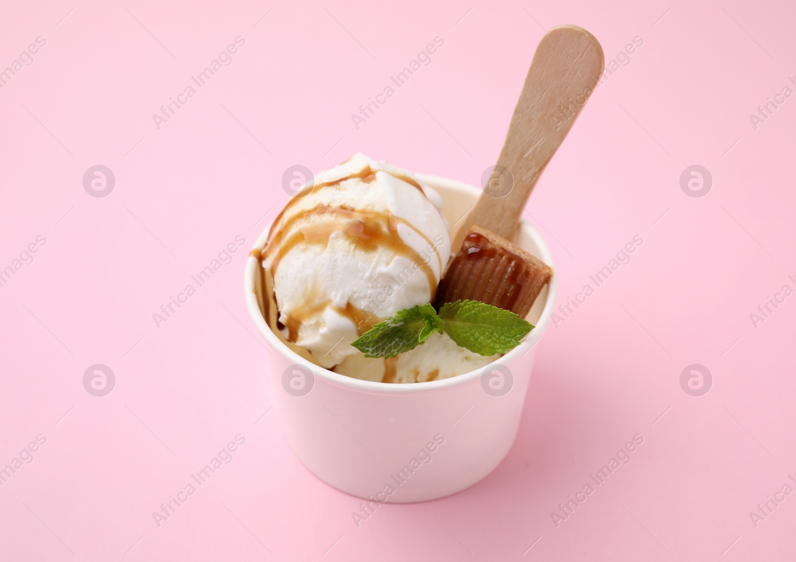 Photo of Tasty ice cream with caramel sauce, mint leaves and candy in paper cup on pink table