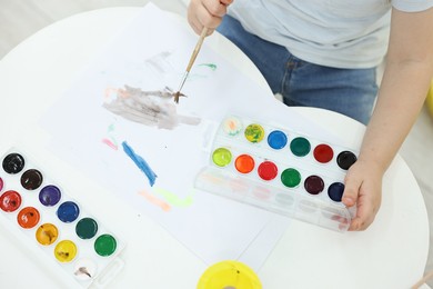 Photo of Little boy painting with brush and watercolor at white table, closeup