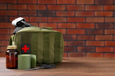 Photo of Military first aid kit, tourniquet, pills and elastic bandage on wooden table against brick wall, space for text