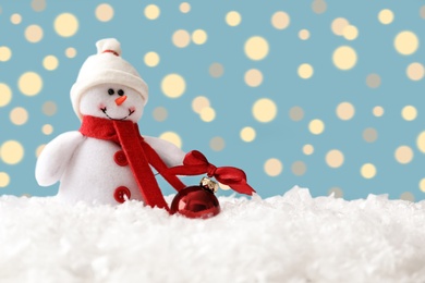 Photo of Snowman toy and Christmas ball on snow against blurred festive lights. Space for text
