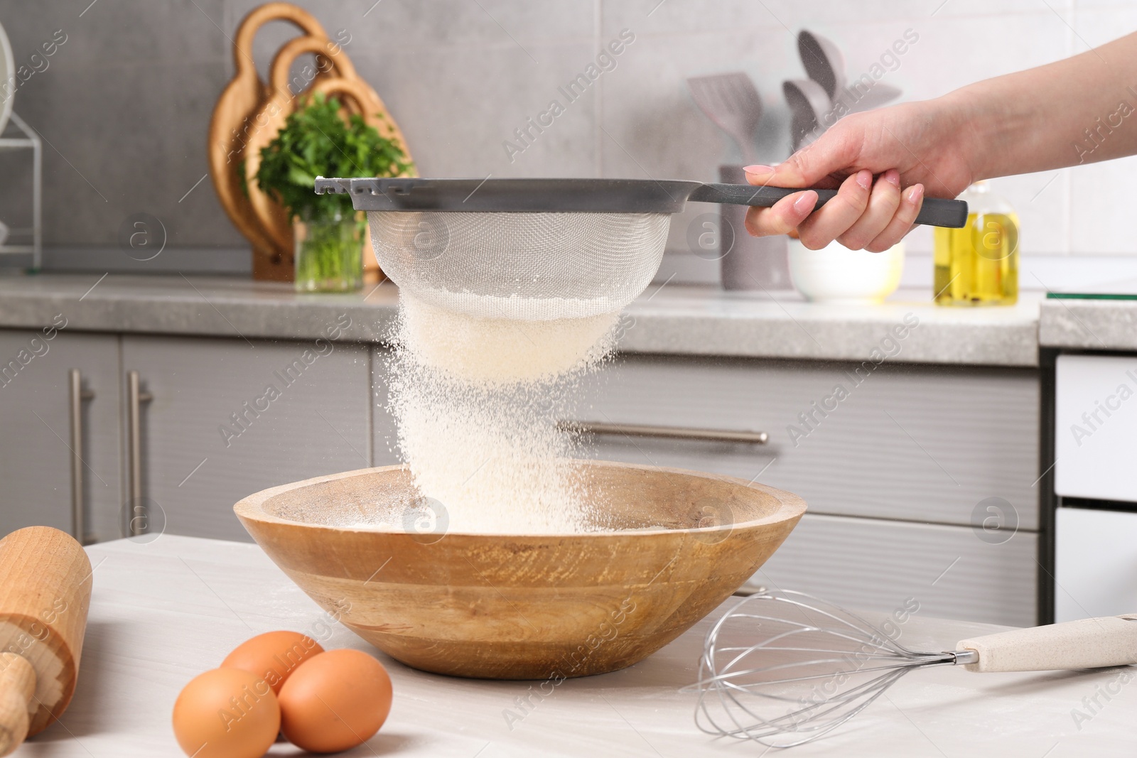 Photo of Woman sieving flour into bowl at white wooden table in kitchen, closeup