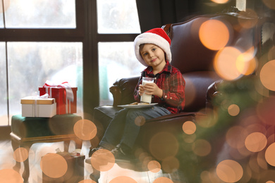 Photo of Little boy in Santa Claus cap with milk and cookies near window indoors. Christmas holiday