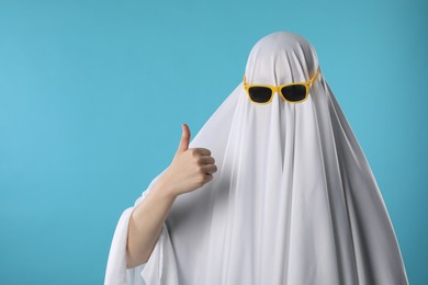 Stylish ghost. Woman covered with white sheet in sunglasses showing thumbs up on light blue background, space for text