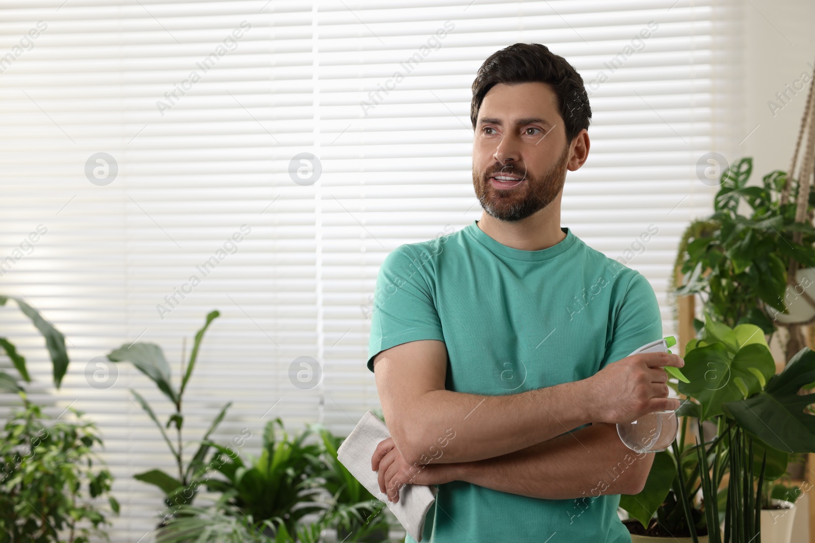 Photo of Portrait of handsome man with spray bottle and cloth near houseplants indoors. Space for text