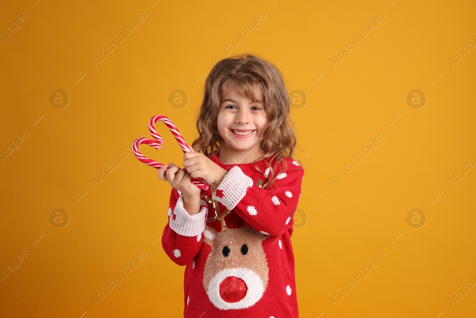 Photo of Cute little girl in Christmas sweater making heart shape with candy canes against orange background