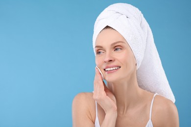 Photo of Beautiful woman in terry towel removing makeup with cotton pad on light blue background, space for text