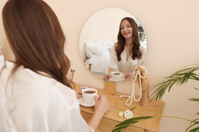 Happy bride with cup of tea near mirror at dressing table. Wedding day