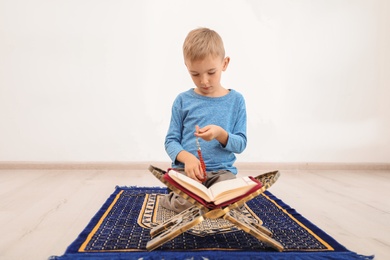 Little Muslim boy with misbaha and Koran praying on rug indoors