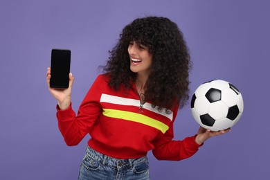 Photo of Happy fan holding soccer ball and showing smartphone on violet background