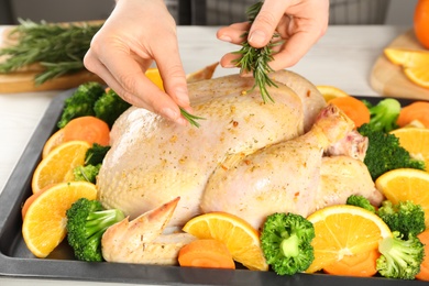 Woman adding rosemary to chicken with oranges and vegetables at table in kitchen, closeup
