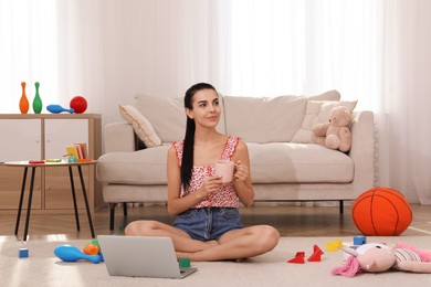 Photo of Happy young mother with cup of coffee and laptop resting in messy living room