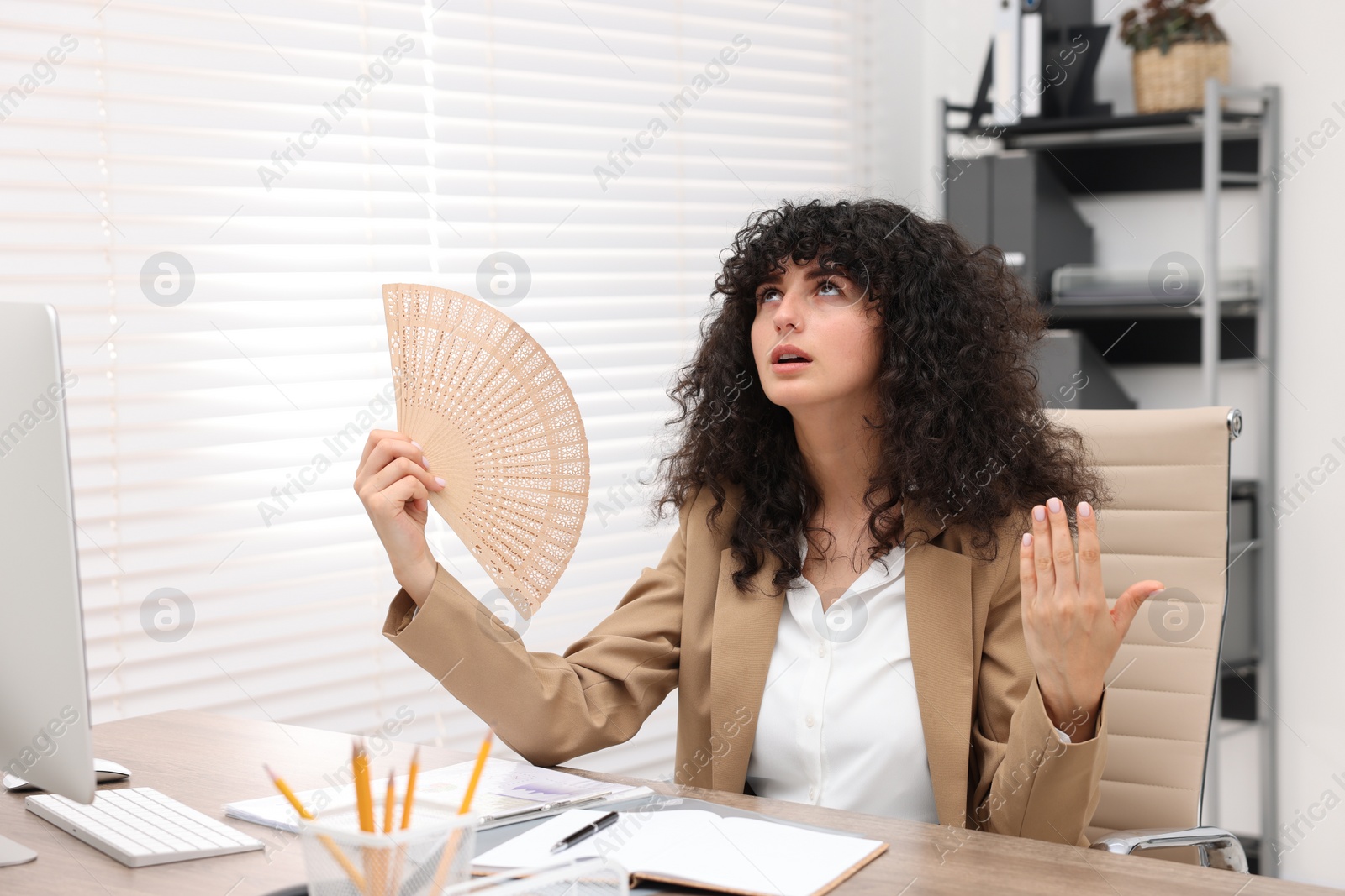 Photo of Young businesswoman waving hand fan to cool herself at table in office