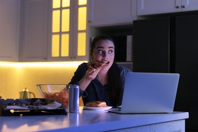 Young woman eating pizza while using laptop in kitchen at night. Bad habit