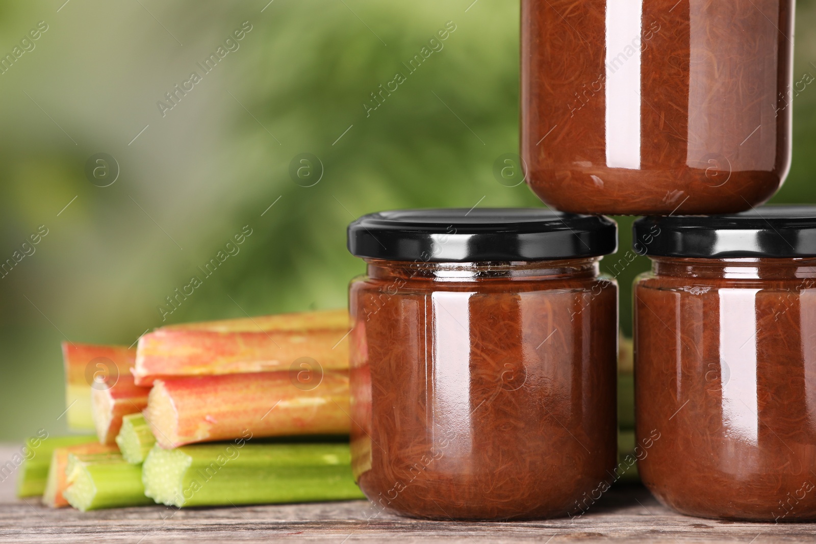 Photo of Jars of tasty rhubarb jam and stalks on wooden table, closeup