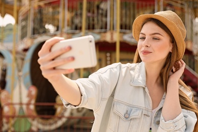 Photo of Young pretty woman taking selfie near carousel in amusement park