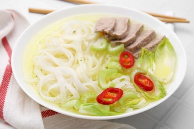 Photo of Bowl of delicious rice noodle soup with celery and meat on white tiled table, closeup