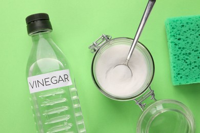 Photo of Eco friendly natural cleaners. Vinegar in bottle, sponge and jar of soda on green background, flat lay