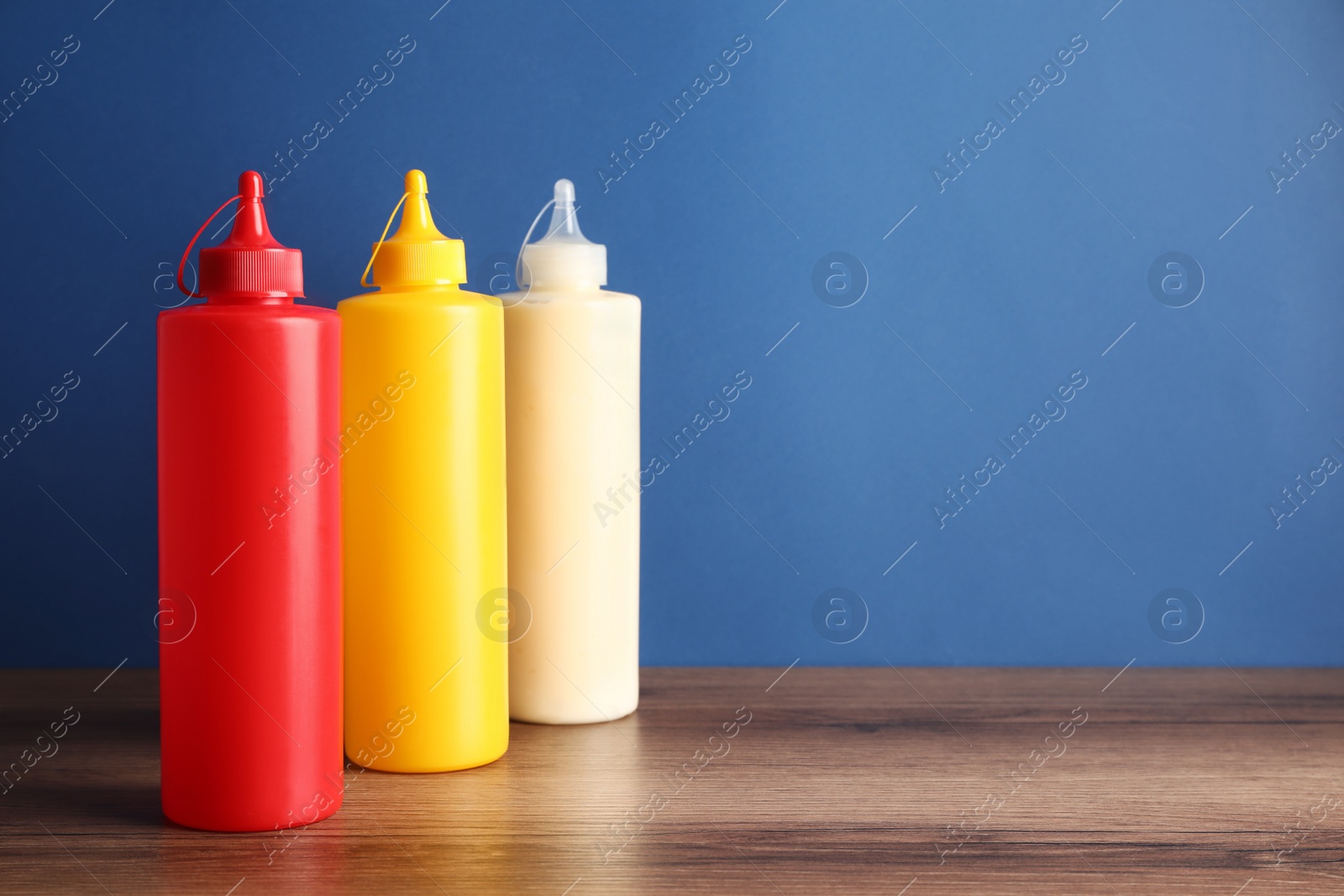 Photo of Ketchup, mustard and mayonnaise in squeeze bottles on wooden table against blue background, space for text