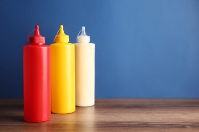Photo of Ketchup, mustard and mayonnaise in squeeze bottles on wooden table against blue background, space for text