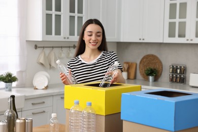 Photo of Garbage sorting. Smiling woman throwing plastic bottles into cardboard box in kitchen