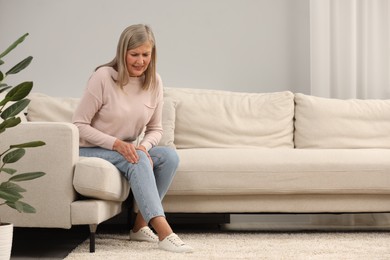 Photo of Mature woman suffering from knee pain on sofa at home. Rheumatism symptom