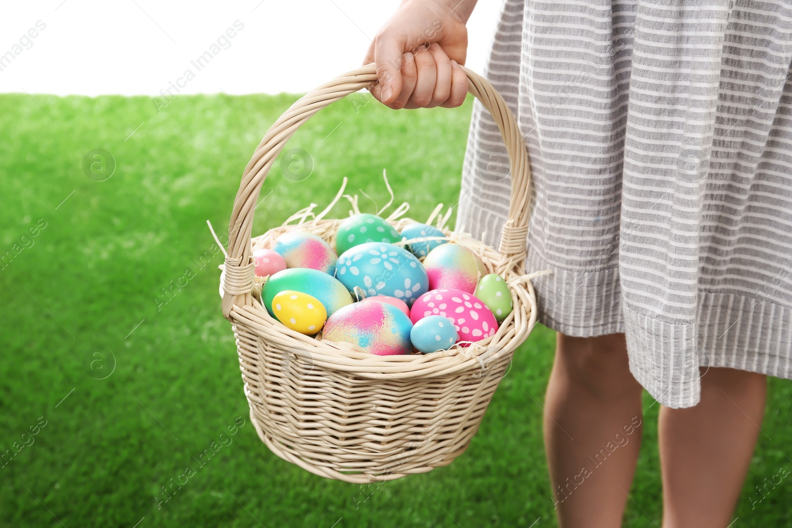 Photo of Little girl with basket full of Easter eggs on green grass against white background, closeup