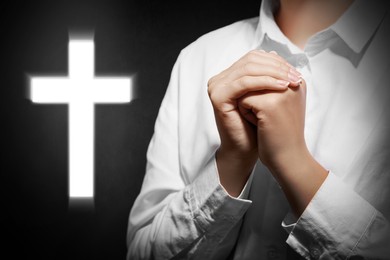 Image of Christian cross and woman holding hands clasped while praying against black background, closeup