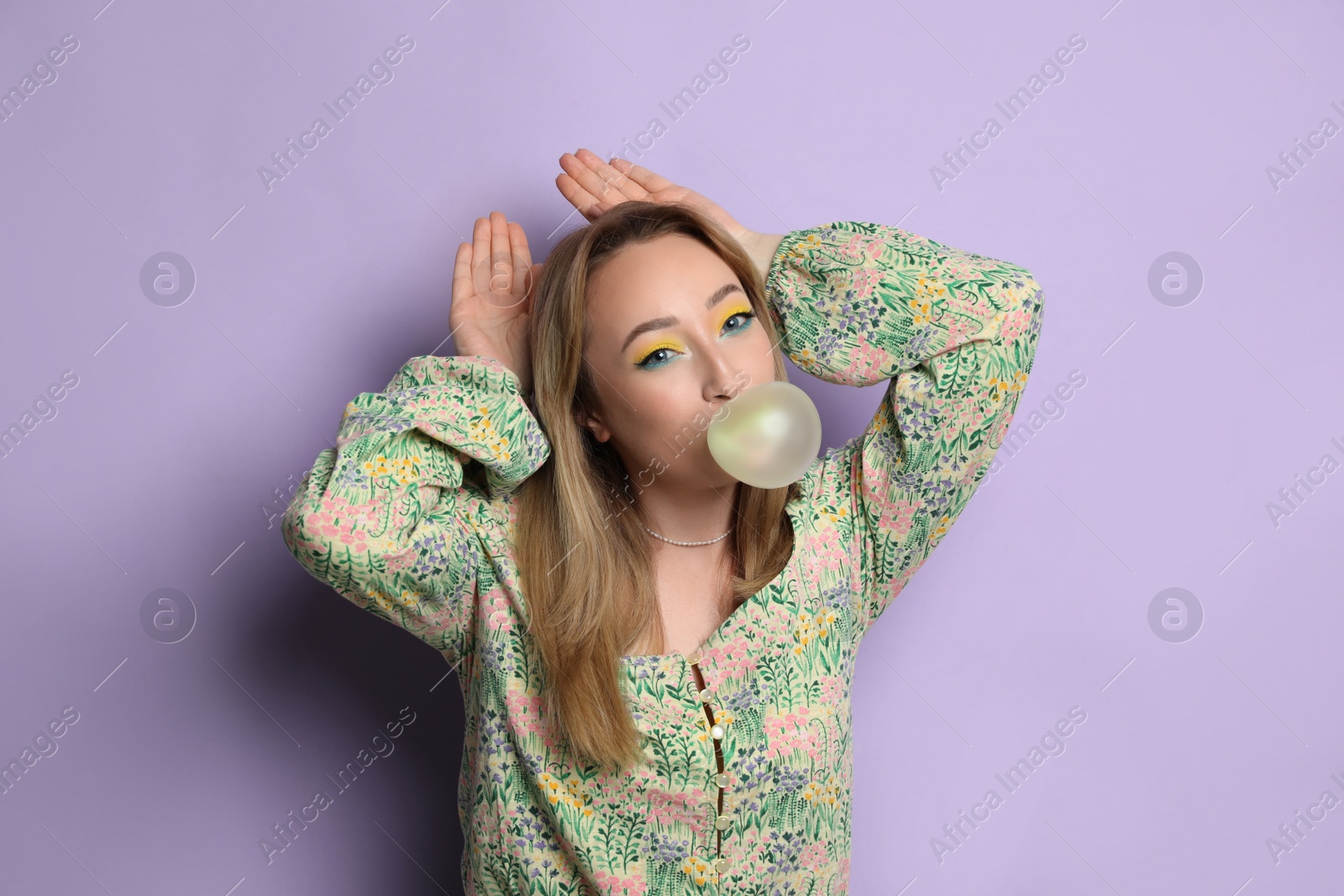 Photo of Fashionable young woman with bright makeup blowing bubblegum on lilac background