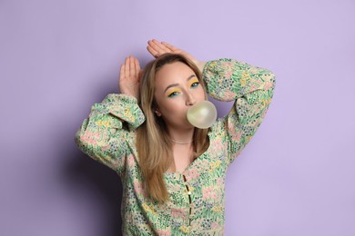 Fashionable young woman with bright makeup blowing bubblegum on lilac background