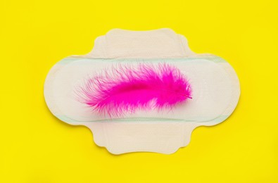 Photo of Menstrual pad with pink feather on yellow background, top view