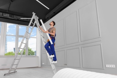 Photo of Electrician in uniform with ceiling lamp on metal ladder indoors. Space for text