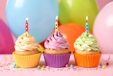 Photo of Birthday cupcakes with burning candles and sprinkles on pink table against color balloons, closeup