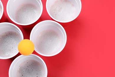 Plastic cups and ball on red background, flat lay with space for text. Beer pong game