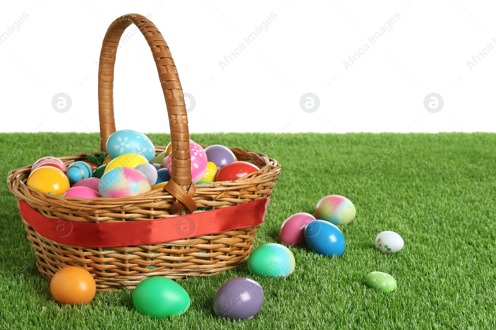 Photo of Wicker basket with Easter eggs on green grass against white background. Space for text