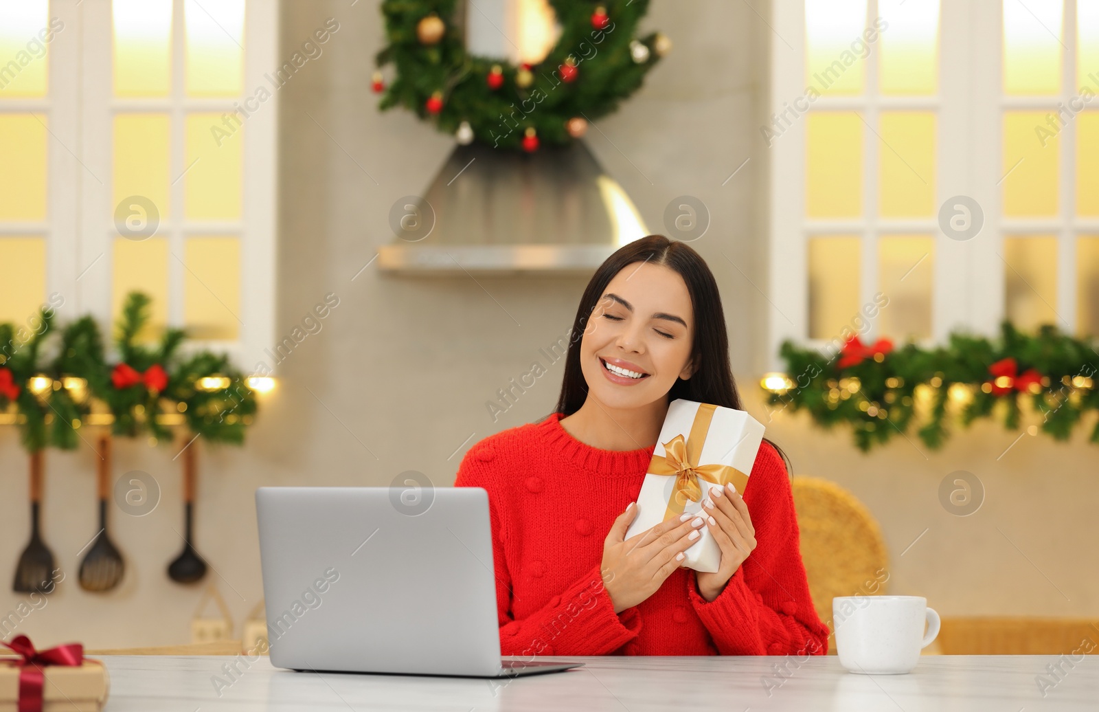 Photo of Celebrating Christmas online with exchanged by mail presents. Smiling woman with gift box during video call on laptop at home