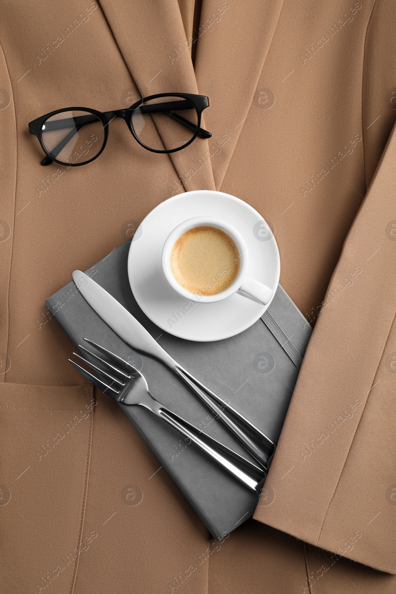 Photo of Business lunch concept. Cup of coffee, light gray notebook and cutlery on beige jacket, flat lay