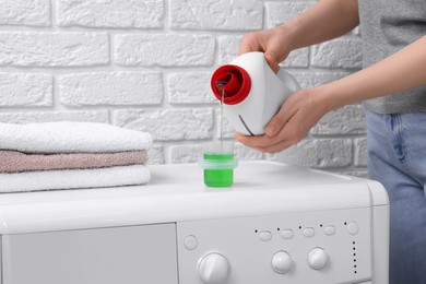 Photo of Woman pouring fabric softener from bottle into cap on washing machine near white brick wall, closeup