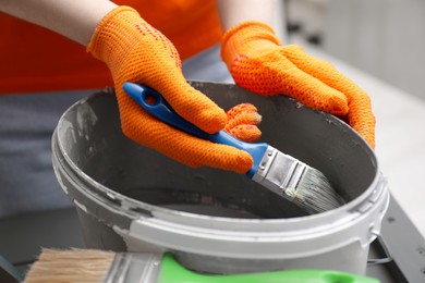 Photo of Woman dipping brush into bucket of grey paint indoors, closeup