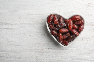 Photo of Heart shaped bowl with sweet dried date fruits on wooden background, top view. Space for text