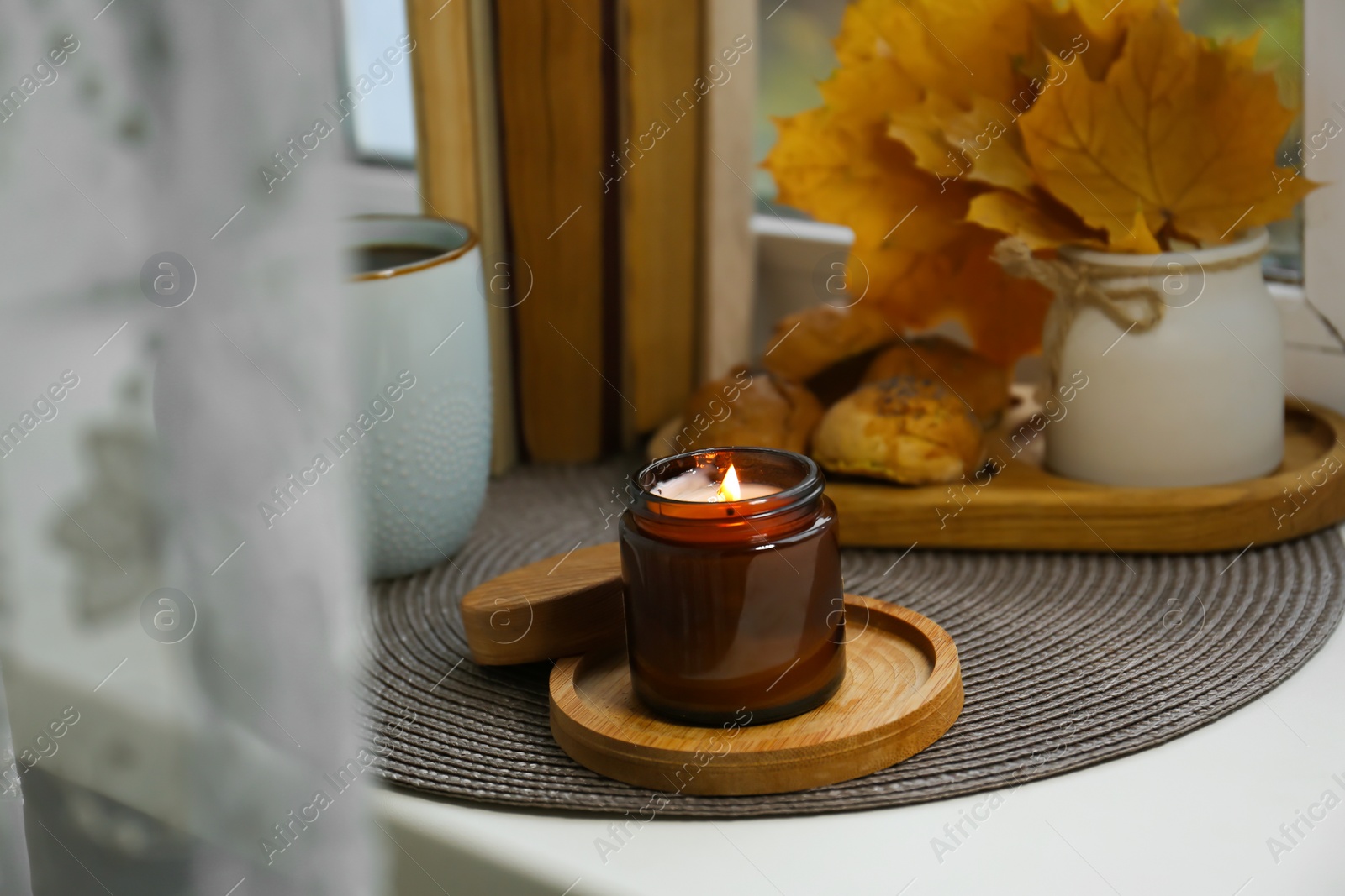 Photo of Burning aromatic candle near cup with hot drink, books, cookies and leaves on wicker mat. Autumn coziness
