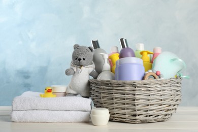 Photo of Wicker basket with different baby cosmetic products, towel and toys on white wooden table