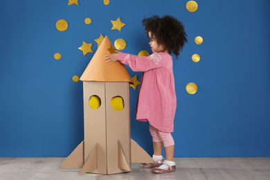 Cute African American child playing with cardboard rocket near blue wall