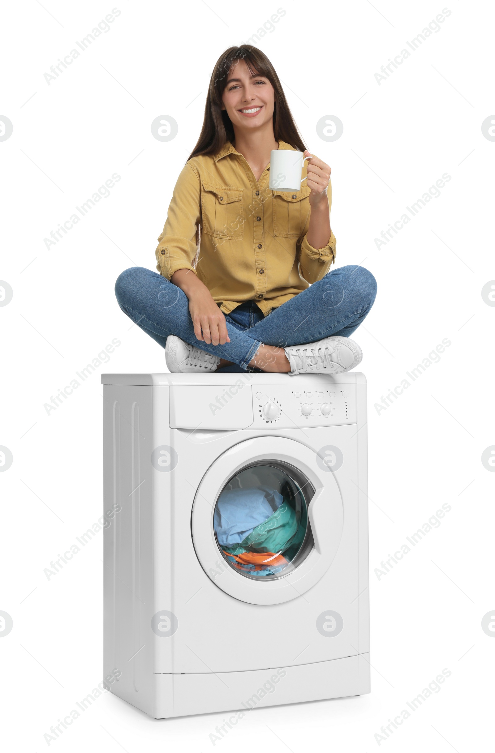 Photo of Beautiful woman with cup of drink sitting on washing machine against white background