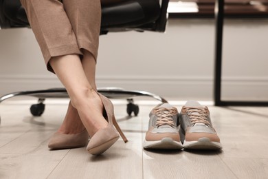 Photo of Comfortable sneakers on floor near woman wearing stylish high heeled shoes in office, closeup