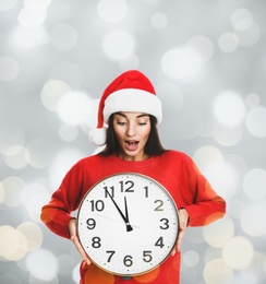 Image of New Year countdown. Surprised woman in Santa hat holding clock on light background