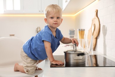 Photo of Curious little boy playing with saucepan on electric stove in kitchen